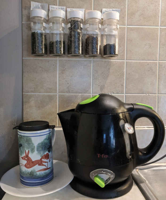 A photograph of an electric T-Fal tea kettle with inserted analogue thermometer, next to a ceramic mug decorated with a fox, containing a black Finum tea filter, all underneath 5 spice jars filled with kukicha and oolong tea, and sitting on a kitchen counter. Electric tea kettle, tea mug, and filter