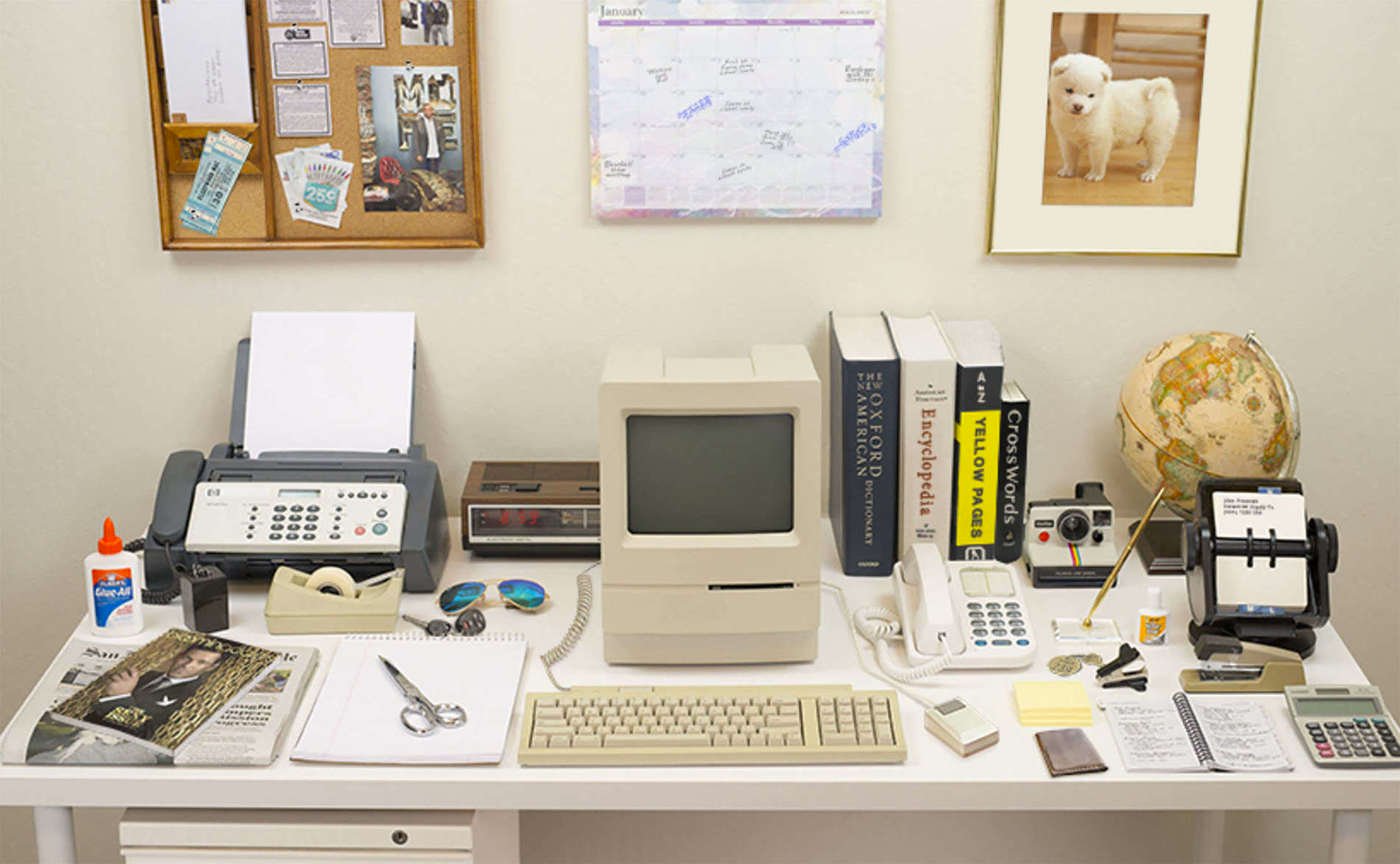 Photograph of a 1980s-vintage desktop, created by the Harvard Innovation Labs in 2015 to demonstrate 'the evolution of the desk', which is cluttered with objects which have since been replaced by computer software/hardware/the Internet. Objects include white glue, saran tape, magazines, newspapers, pencil sharpener, fax machine, car keys, scissors, notepad, Macintosh, radio clock, Oxford English Dictionary, encyclopedia, Yellow Pages, crosswords collection, phone, Post-Its, wallet, camera, fancy pen, whiteout fluid, calculator, Rolodex, globe, cork board, and other objects. The 1980s Desktop